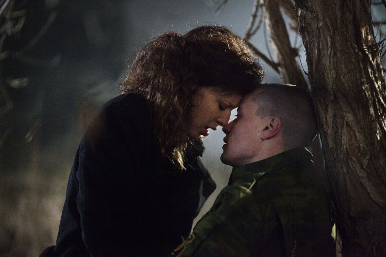 Kobieta i mężczyzna w wojskowym mundurze siedzą nocą blisko siebie pod drzewem, stykają się czołami, a na ich twarzach malują się emocje.