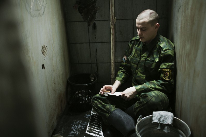 Osoba w wojskowym kamuflażu siedzi na wąskiej ławce w ciemnym, małym, brudnym pokoju, czytając list. W pobliżu znajdują się metalowe wiadra i kosz na śmieci, a ściany są poplamione i puste.