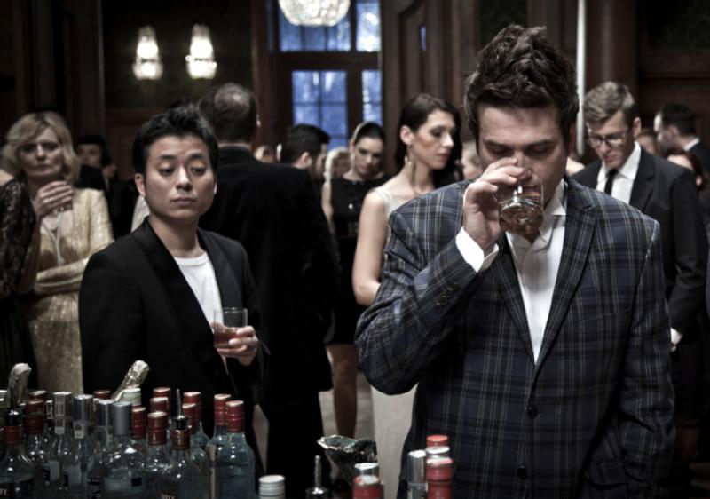 Grupa ludzi ubranych w formalne stroje zbiera się na eleganckim przyjęciu. Dwie osoby stoją na pierwszym planie przy barze z butelkami, jedna pije ze szklanki, a druga patrzy w stronę aparatu.