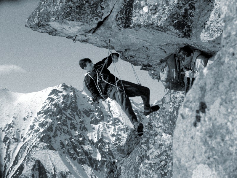 Dwóch wspinaczy wspina się na stromą, skalistą ścianę klifu z pokrytymi śniegiem górami w tle. Jeden z nich wisi na linach, a drugi jest częściowo ukryty za półką skalną. Obaj mają na sobie sprzęt wspinaczkowy.