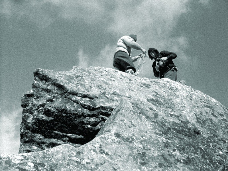Dwie osoby w ubraniach outdoorowych stoją na skalistym szczycie, poprawiając liny wspinaczkowe lub sprzęt. Zdjęcie zostało zrobione od dołu, podkreślając dużą skałę i częściowo zachmurzone niebo nad nią.
