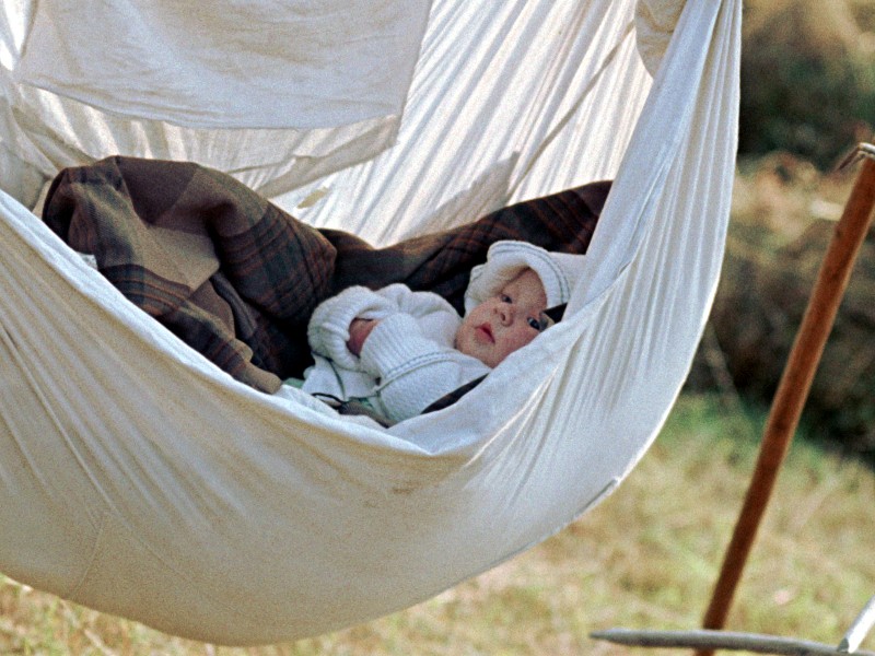Dziecko zawinięte w biały sweter i czapeczkę leży na zewnątrz w białym hamaku z tkaniny, częściowo przykryte kratą. Scena jest skąpana w miękkim, naturalnym świetle.