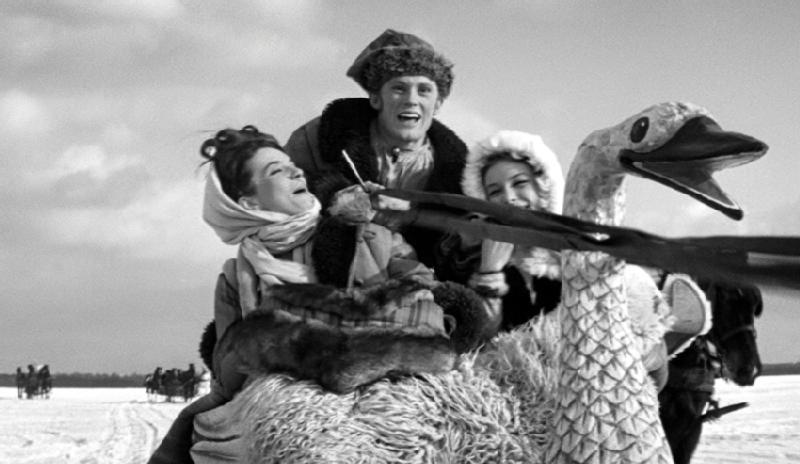 Trzy osoby ubrane w zimowe ubrania śmieją się radośnie, jadąc na dużych saniach w kształcie łabędzia po zaśnieżonym krajobrazie, na tle jasnego nieba.