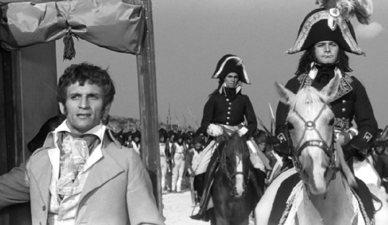 Mężczyzna w stroju z XIX wieku stoi na pierwszym planie, a za nim dwóch mężczyzn w wyszukanych mundurach wojskowych jedzie konno, prowadząc grupę żołnierzy. Obraz jest czarno-biały.