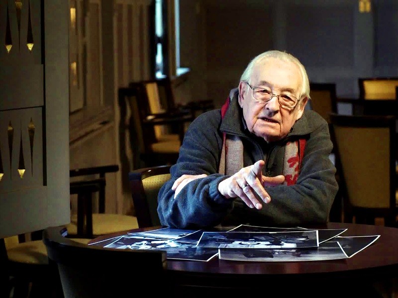 Starszy mężczyzna w okularach siedzi przy stole pokrytym czarno-białymi fotografiami, gestykulując ręką. Pomieszczenie jest delikatnie oświetlone pustymi krzesłami i oknem w tle.