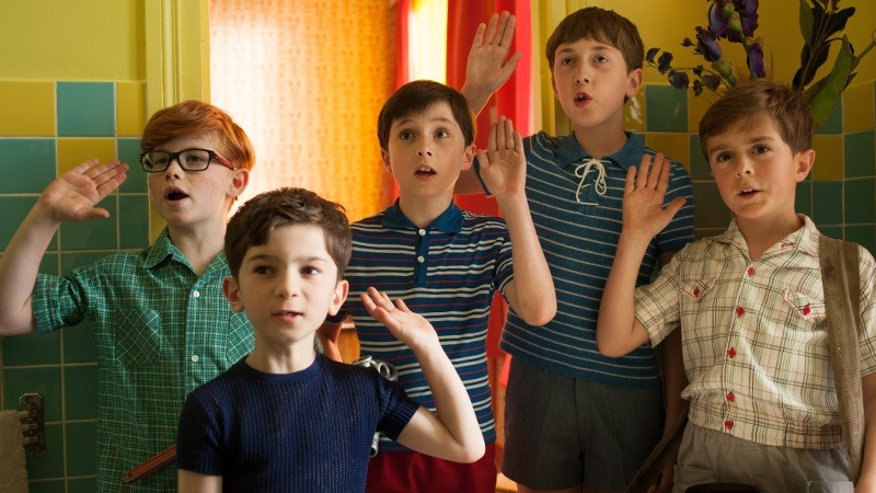 Pięciu chłopców stoi w pomieszczeniu z uniesionymi prawymi rękami, jakby składali przysięgę. Noszą kolorowe ubrania w stylu retro i wyglądają na skupionych. Tło stanowią kafelkowe żółte i zielone ściany z jasnym oświetleniem.