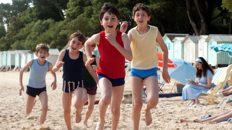 Czwórka dzieci w bluzkach bez rękawów i krótkich spodenkach biegnie w stronę kamery po piaszczystej plaży, z domkami plażowymi i drzewami w tle. Kobieta w niebieskim siedzi na ręczniku i czyta. Scena jest jasna i pełna energii.