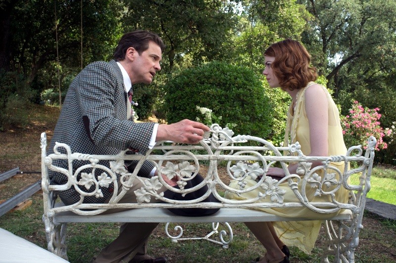 Mężczyzna w kraciastym garniturze i kobieta w żółtej sukience siedzą naprzeciwko siebie na białej ozdobnej ławce w ogrodzie, pogrążeni w poważnej rozmowie. Otacza ich bujna zieleń.