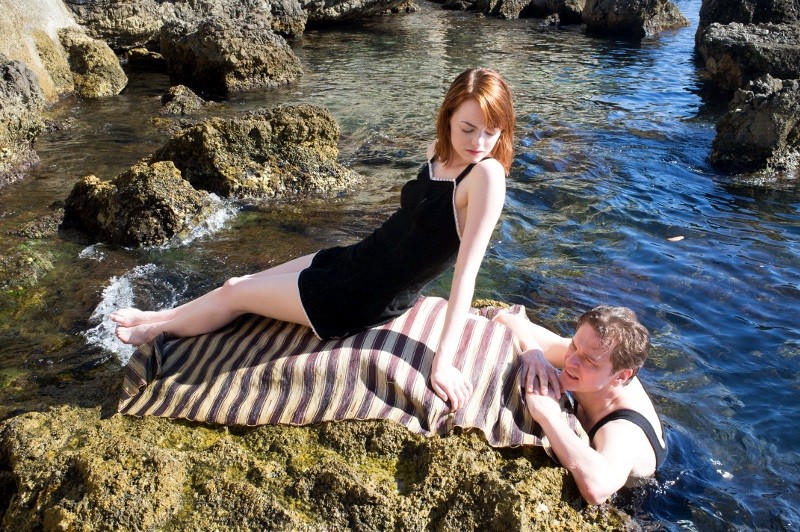 Kobieta w czarnej sukience wyleguje się na pasiastym kocu rozłożonym na skałach nad wodą, podczas gdy mężczyzna w czarnym topie bez rękawów opiera się o skały, częściowo zanurzony w wodzie, i patrzy na nią.