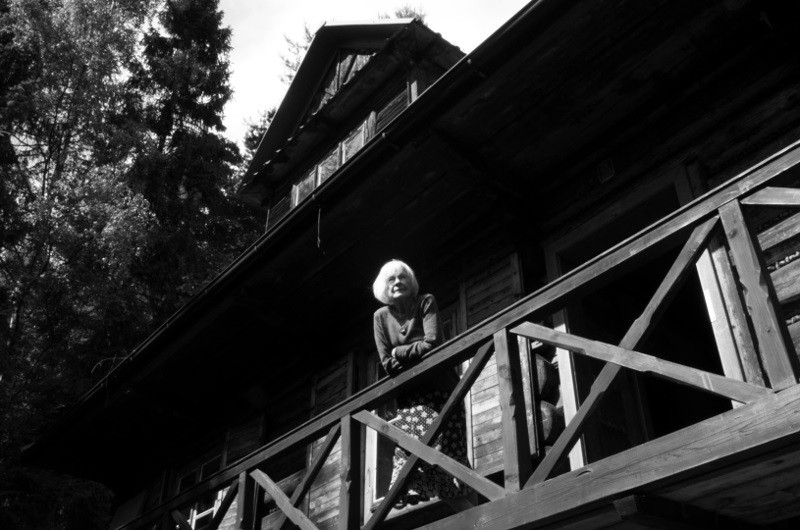 Starsza kobieta o jasnych włosach stoi na drewnianym balkonie rustykalnego, ciemnego domu otoczonego drzewami, patrzy w dół i lekko się uśmiecha. Obraz jest czarno-biały.
