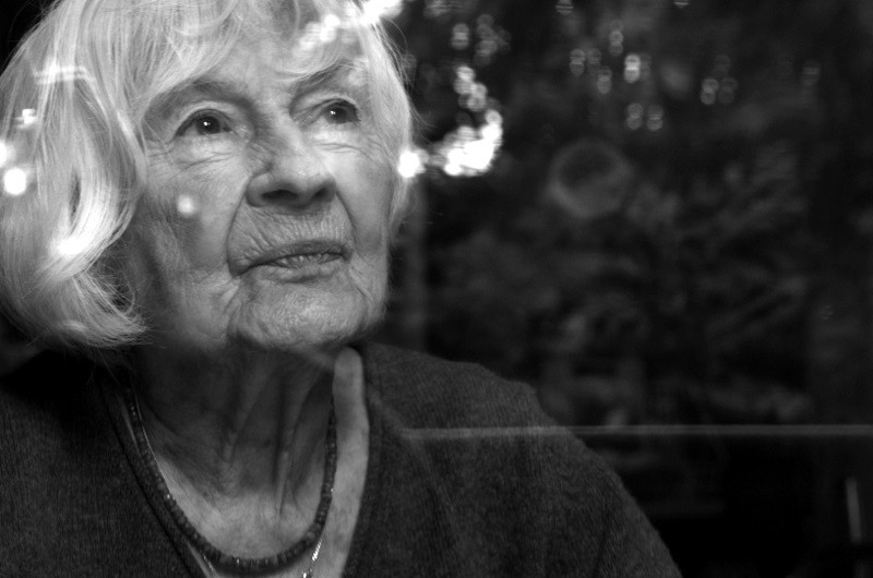 Starsza kobieta z białymi włosami zamyślona spogląda przez okno. Odbicia z zewnątrz delikatnie nakładają się na jej twarz, a ona nosi naszyjnik i sweter. Obraz jest czarno-biały.