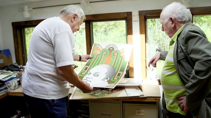 Dwóch starszych mężczyzn stoi w oświetlonym słońcem pokoju, oglądając kolorowe plakaty. Jeden z nich trzyma wydruk przedstawiający stylizowany portret osoby z geometrycznymi wzorami, podczas gdy drugi uważnie go obserwuje. Półki i okno wypełniają tło.