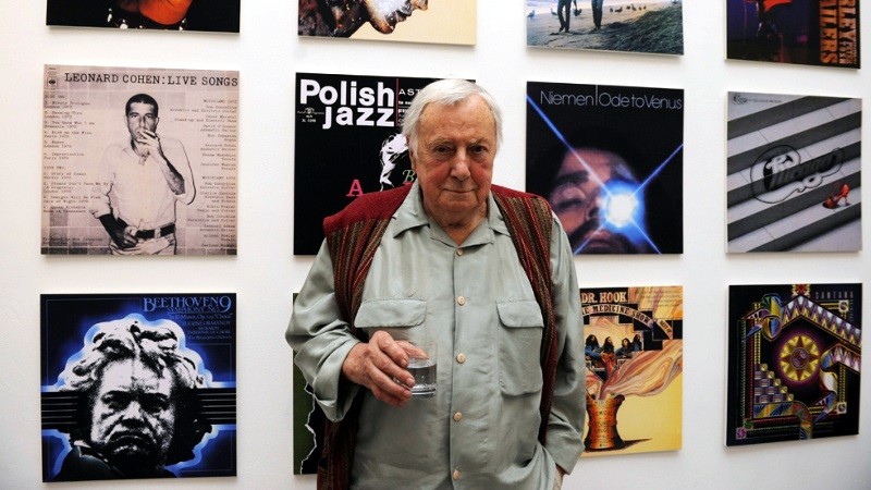 Starszy mężczyzna w jasnej koszuli stoi, trzymając szklankę, przed ścianą pokrytą okładkami różnych albumów muzycznych, w tym jedną z utworami Leonarda Cohena i inną z utworami Beethovena.