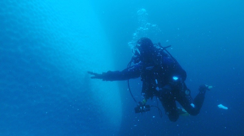 Płetwonurek w pełnym rynsztunku pływa pod wodą w pobliżu dużej, zacienionej, pionowej struktury. Z nurka unoszą się bąbelki, a otaczająca woda jest głęboko niebieska z unoszącymi się cząsteczkami.