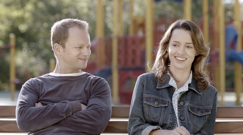 Mężczyzna i kobieta siedzą na ławce na zewnątrz, uśmiechają się i patrzą na siebie. W tle widać rozmazany plac zabaw.
