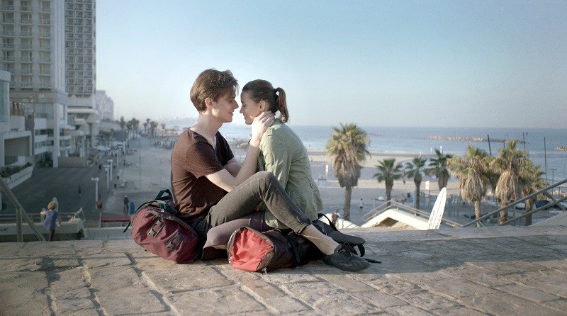 Młoda para siedzi naprzeciwko siebie na kamiennych schodach w pobliżu plaży, dotykając się czołami i trzymając się blisko siebie. Plecaki leżą obok nich, a w tle widać palmy, promenadę i spokojne morze.