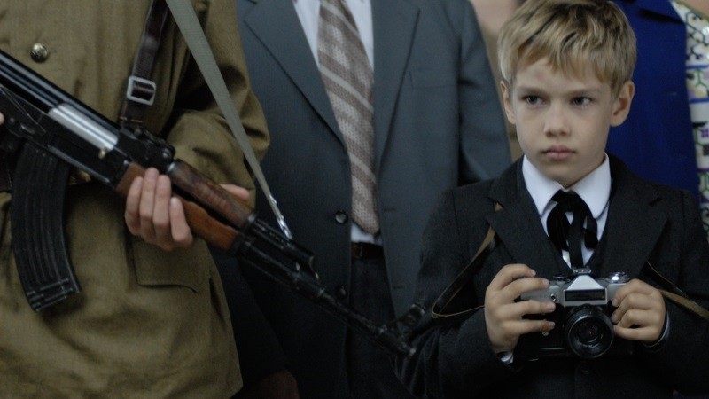 Młody chłopiec w garniturze trzymający aparat fotograficzny stoi obok dorosłego w mundurze wojskowym trzymającego karabin. Chłopiec wygląda poważnie i jest otoczony przez dorosłych w formalnych ubraniach.