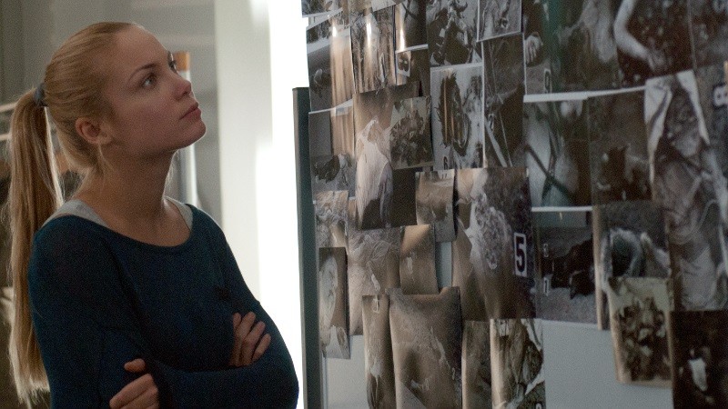 Kobieta z blond włosami związanymi w kucyk stoi ze skrzyżowanymi ramionami i uważnie wpatruje się w ścianę pokrytą czarno-białymi fotografiami i notatkami, jakby analizowała planszę śledczą.