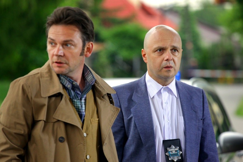 Dwóch mężczyzn, jeden w brązowym płaszczu, a drugi w niebieskim blezerze z policyjną odznaką na szyi, stoi na zewnątrz, patrząc w tym samym kierunku z poważnymi minami. W tle widać drzewa i rozmazany budynek.