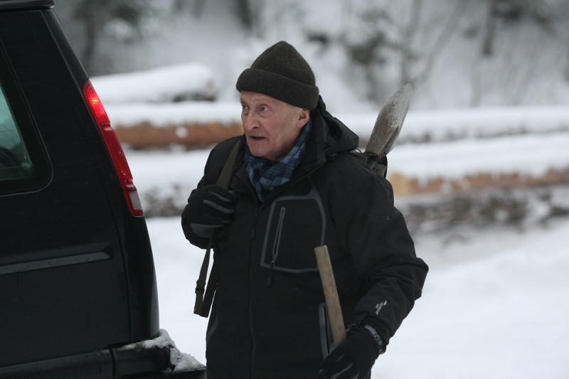 Starszy mężczyzna ubrany w ciemną kurtkę, czapkę i szalik, niesie łopatę na ramieniu i stoi obok czarnego pojazdu na zaśnieżonym terenie.