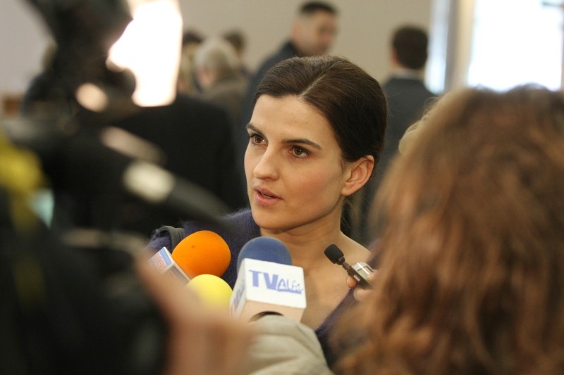 Kobieta o ciemnych włosach przemawia do reporterów, w otoczeniu kilku mikrofonów oznaczonych różnymi logotypami mediów, w zatłoczonym pomieszczeniu, z niewyraźnymi postaciami w tle.