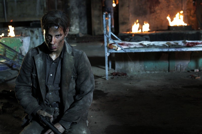 Rozczochrany, ranny mężczyzna w brudnym ubraniu siedzi na podłodze, trzymając broń w ciemnym, zniszczonym pokoju, w tle płoną ogniska i zakrwawione łóżko, co sugeruje scenę pełną przemocy i chaosu.