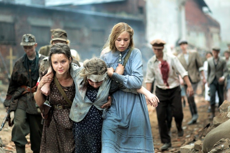 Grupa zrozpaczonych ludzi w staromodnych ubraniach przechodzi przez gruzy; dwie młode kobiety wspierają ranną dziewczynę, podczas gdy inni, niektórzy wyglądający na rannych, podążają za nimi. Scena sugeruje wojnę lub sytuację po katastrofie.