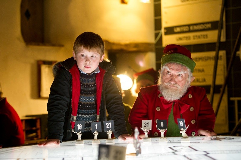 Młody chłopiec w swetrze stoi obok starszego mężczyzny przebranego za elfa, obaj pochylają się nad stołem z ponumerowanymi znacznikami. Sceneria wydaje się świąteczna z dekoracjami świątecznymi w tle.