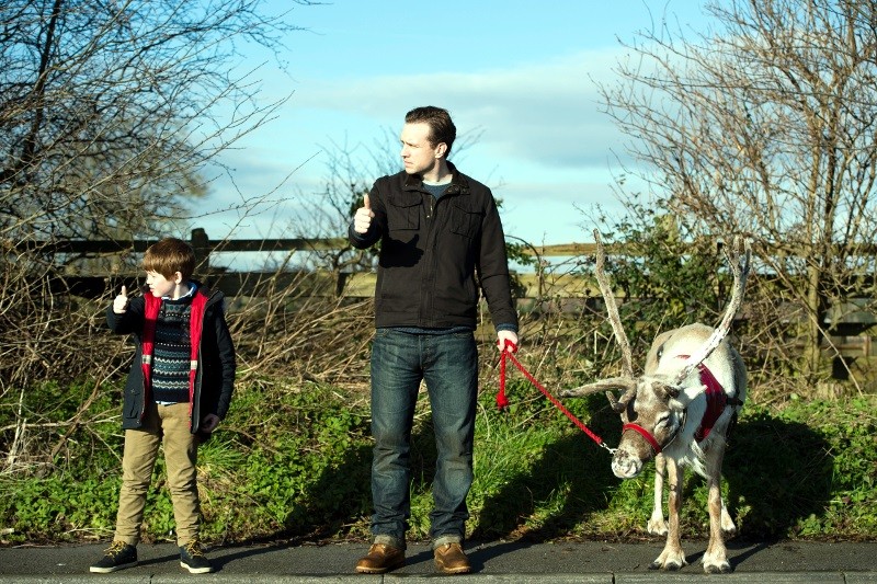 Mężczyzna i chłopiec stoją na zewnątrz na chodniku i podnoszą kciuki, podczas gdy mężczyzna trzyma czerwoną smycz przywiązaną do białego renifera. Nagie drzewa i drewniany płot znajdują się w tle pod błękitnym niebem.