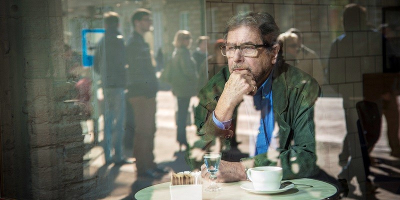 Starszy mężczyzna w okularach siedzi sam przy stoliku kawiarnianym przy oknie, opierając brodę na dłoni i wyglądając na zamyślonego. W szkle widać odbicia ludzi spacerujących na zewnątrz. Na stole stoi filiżanka i szklanka wody.
