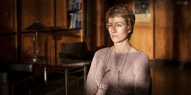 Kobieta z krótkimi włosami siedzi w środku, ubrana w jasnofioletowy sweter i okulary wiszące na szyi. Wydaje się zamyślona, a pokój za nią ma drewniane panele, półki i stół z lampą.