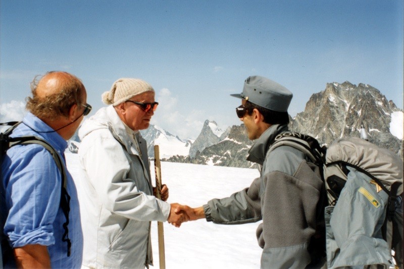 Trzech mężczyzn stoi na ośnieżonym górskim krajobrazie. Dwóch ściska sobie dłonie; jeden nosi szary mundur i okulary przeciwsłoneczne, drugi beżową czapkę i białą kurtkę. Trzeci mężczyzna, ubrany na niebiesko, przygląda się. W tle wznoszą się skaliste szczyty.