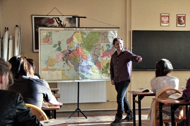 Nauczyciel stoi przy dużej mapie Europy, wskazując i rozmawiając z uczniami siedzącymi przy ławkach w klasie. W klasie jest tablica, a do środka wpada światło słoneczne.