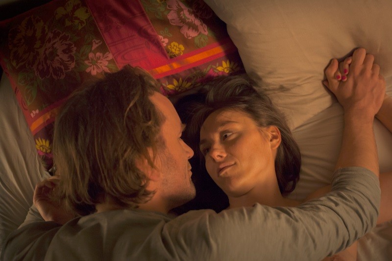 Mężczyzna i kobieta leżą na łóżku naprzeciwko siebie, trzymając się za ręce. Kobieta patrzy mężczyźnie w oczy, które są częściowo przykryte kocem. Za nimi znajduje się kolorowa poduszka. Scena wydaje się intymna i czuła.