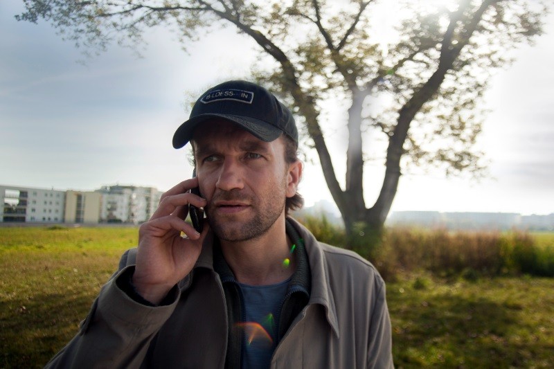 Mężczyzna ubrany w czarną czapkę i beżową kurtkę stoi na zewnątrz w słoneczny dzień, rozmawiając przez telefon komórkowy, w tle widać duże drzewo i trawiaste pole.