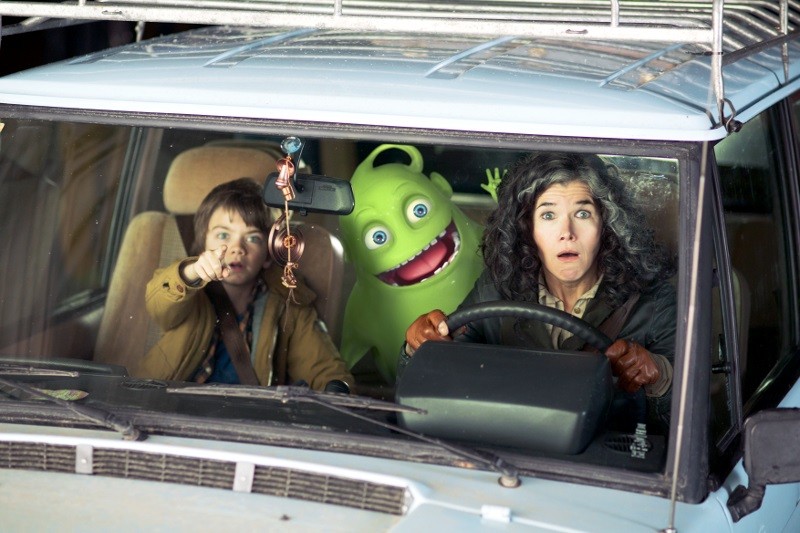Zaskoczona kobieta prowadzi samochód, w którym młody chłopiec wskazuje przed siebie z siedzenia pasażera, a między nimi siedzi uśmiechnięty zielony potwór z kreskówki. Wszyscy troje wydają się zaskoczeni, gdy patrzą przez przednią szybę.