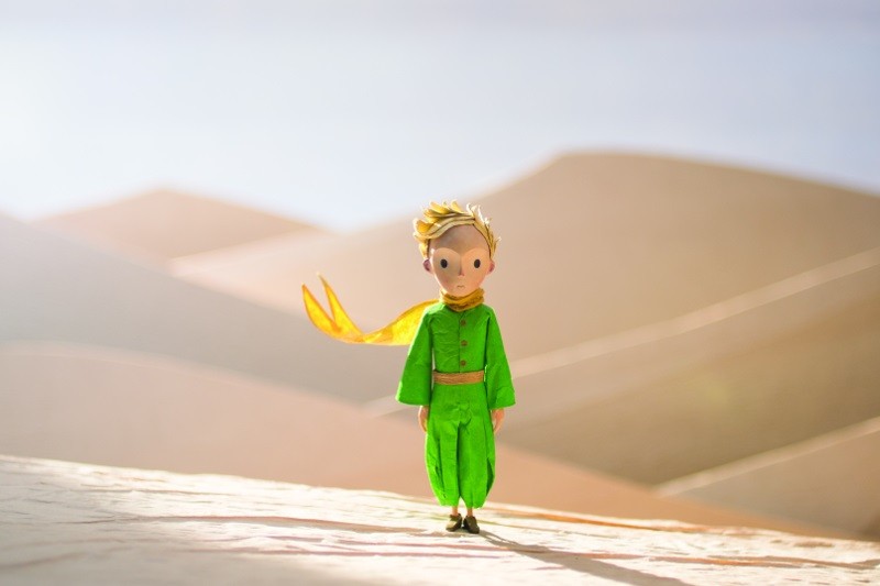 Mała figurka chłopca w jaskrawozielonym stroju i żółtym szaliku stoi samotnie na bladych wydmach pod jasnym niebem.