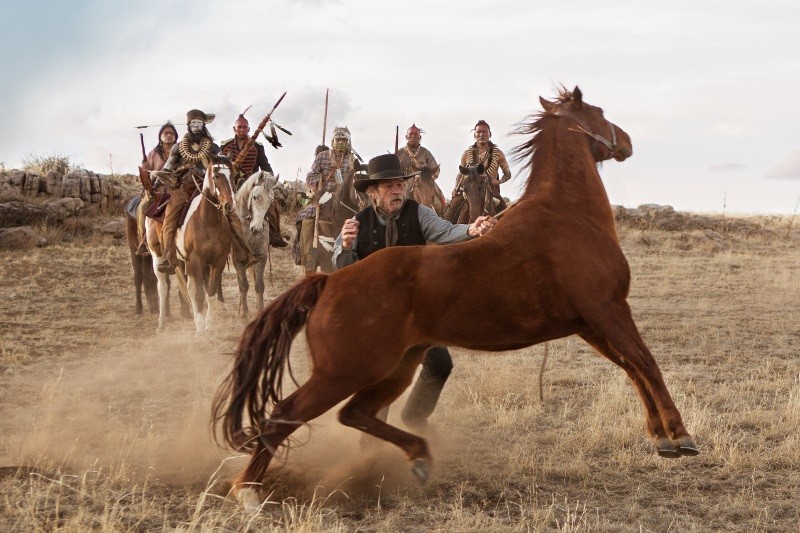 Mężczyzna w kowbojskim kapeluszu próbuje opanować stającego dęba brązowego konia na suchym polu trawy, podczas gdy grupa konnych wojowników indiańskich z włóczniami i piórami zbliża się od tyłu.