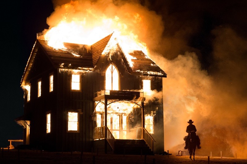 Duży dom jest objęty płomieniami w nocy, ogień bucha z dachu i okien. Na pierwszym planie osoba na koniu obserwuje płonący dom, którego sylwetka odcina się od płomieni.