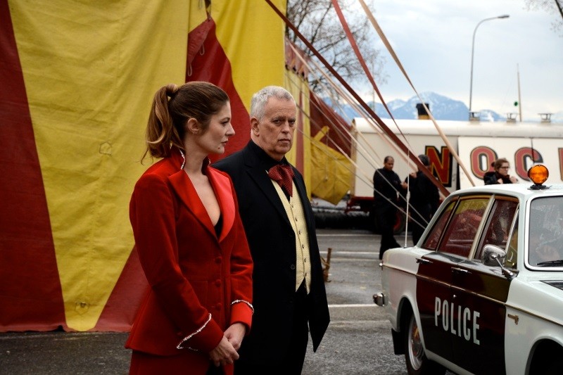 Kobieta w czerwonym garniturze i mężczyzna w garniturze stoją obok zabytkowego radiowozu w pobliżu dużego, żółto-czerwonego namiotu cyrkowego. Scena wydaje się napięta i poważna.