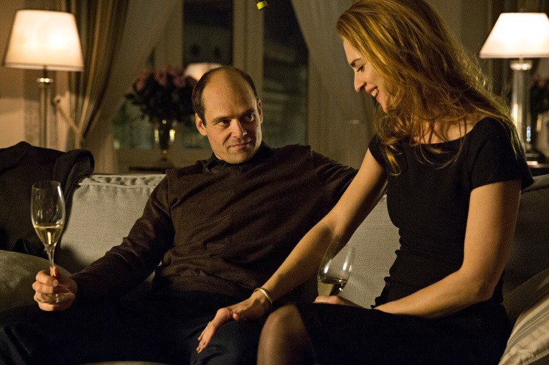 Mężczyzna i kobieta siedzą blisko siebie na kanapie w ciepło oświetlonym pokoju, każdy trzymający kieliszek wina. Uśmiechają się i wydają się cieszyć swoim towarzystwem.