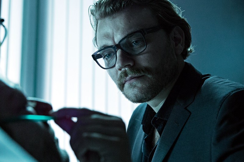 Mężczyzna z falowanymi włosami, brodą i okularami, ubrany w garnitur, uważnie przygląda się komuś, trzymając w górze parę okularów, w słabo oświetlonym pomieszczeniu z pionowymi żaluzjami w tle.