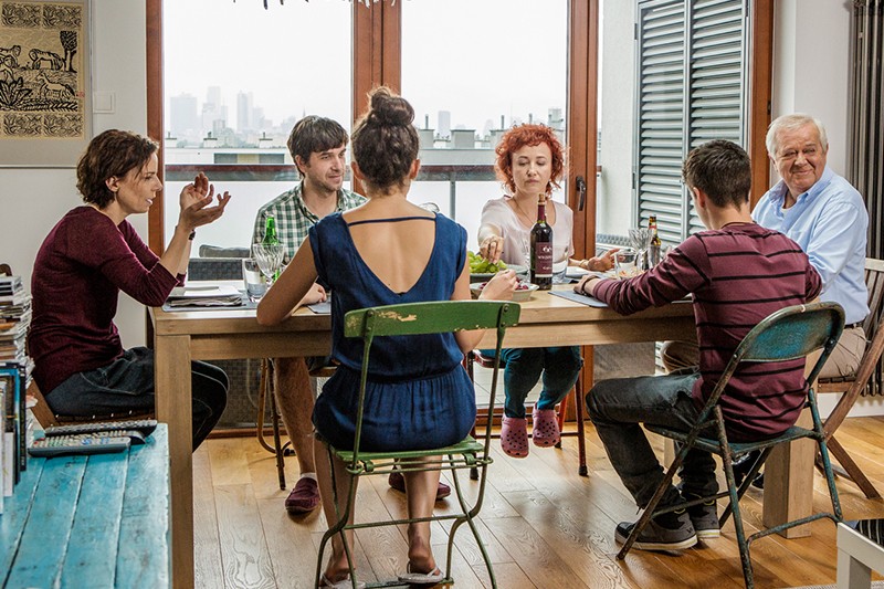 Sześć osób w różnym wieku siedzi przy stole jadalnym w przytulnym, nasłonecznionym pokoju. Na stole leżą jedzenie, napoje i papiery, a grupa wydaje się być zajęta rozmową. Przez okna widać panoramę miasta.