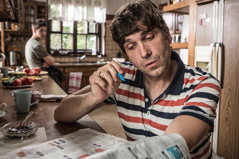 Mężczyzna w pasiastej koszuli siedzi przy kuchennym blacie, czyta gazetę i trzyma długopis. Inny mężczyzna jest w tle przy oknie, patrząc w jego stronę. Kuchnia ma drewniane szafki i różne przedmioty na blacie.