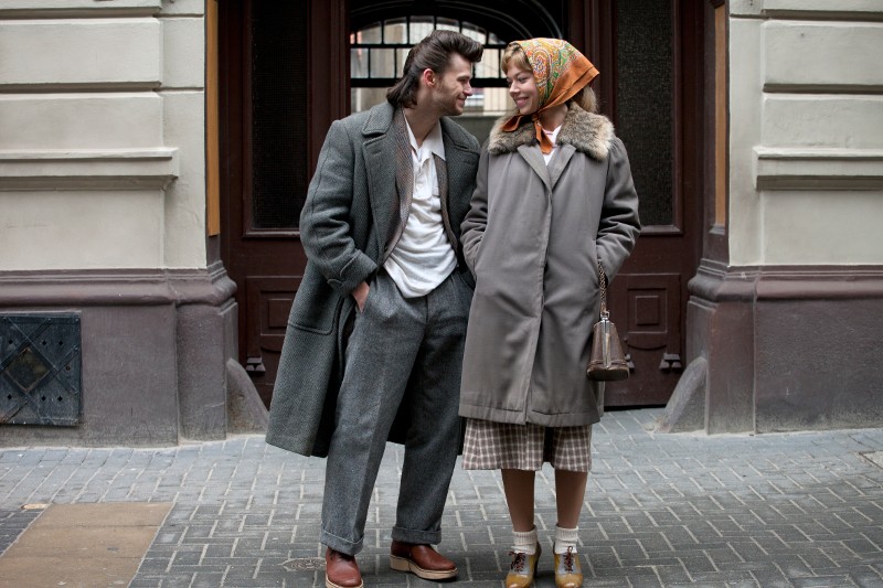 Mężczyzna i kobieta ubrani w płaszcze w stylu vintage, stojący na brukowanej ulicy przed budynkiem. Kobieta ma kolorową chustę na głowie, a oboje się do siebie uśmiechają.