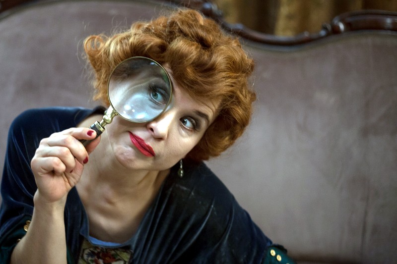 Kobieta z kręconymi rudymi włosami trzyma lupę przy jednym oku, przez co jej oko wydaje się większe. Nosi ciemnoniebieski top i jaskrawoczerwoną szminkę i patrzy figlarnie w kamerę.