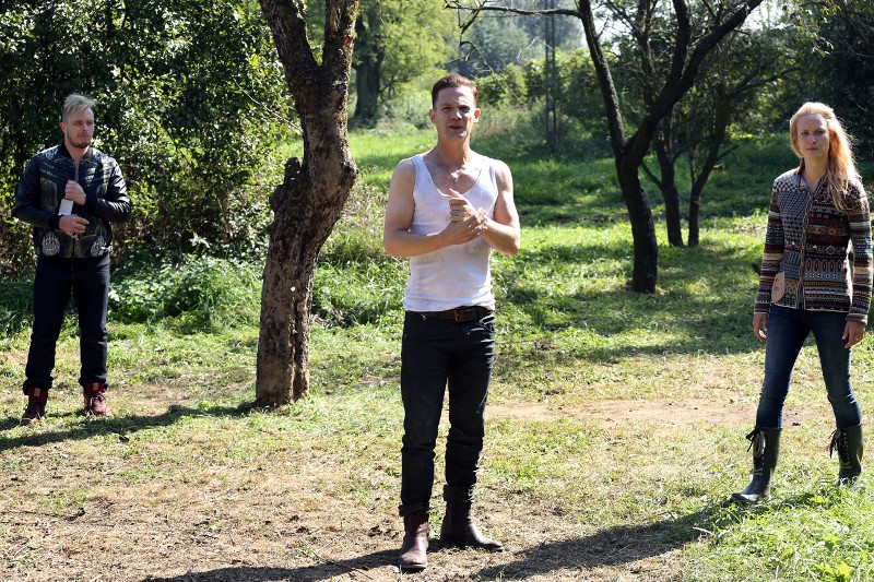 Trzy osoby stoją osobno w rozświetlonym słońcem, trawiastym parku otoczonym drzewami. Mężczyzna w środku ma na sobie biały podkoszulek, kobieta po prawej ma na sobie buty i wzorzysty sweter, a mężczyzna po lewej ma na sobie czarną kurtkę.