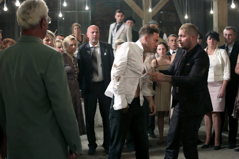 Grupa osób w formalnych strojach obserwuje, jak dwóch mężczyzn, jeden w podartej białej koszuli, kłóci się lub walczy w centrum pokoju oświetlonego wiszącymi żarówkami.