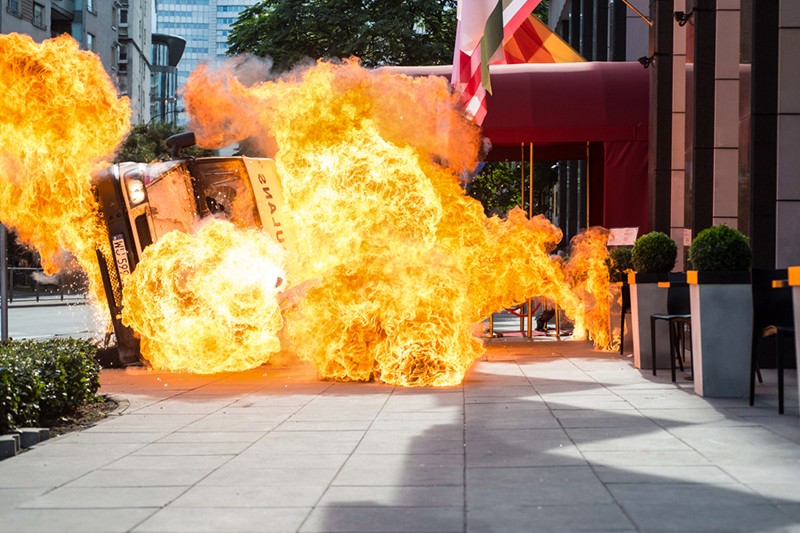 Ogromna kula ognia pochłania pojazd, który najprawdopodobniej przewraca się na miejskim chodniku. W pobliżu wejścia do budynku szybko rozprzestrzeniają się płomienie i dym.