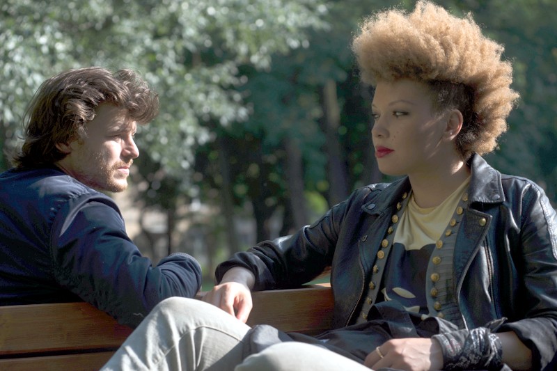 Dwie osoby siedzą na ławce w parku i prowadzą poważną rozmowę. Osoba po lewej ma falowane włosy i brodę, podczas gdy osoba po prawej ma dużą, kręconą fryzurę i nosi nabijaną ćwiekami kurtkę. W tle widać drzewa.